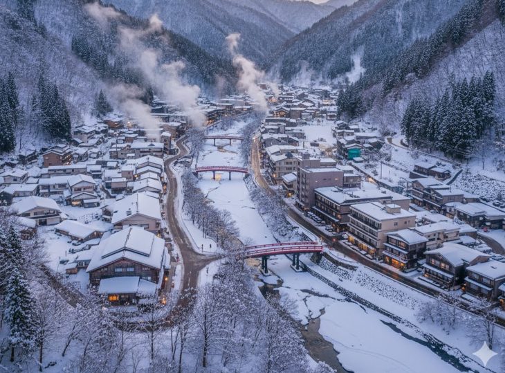 雪に覆われた山々と、その手前に広がる四万温泉街の全景。朝焼けや夕焼けで空が少し色づき、冬の始まりの静けさを感じる。